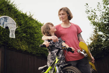 Vater und Sohn mit Fahrrad und Skateboard im Hof gegen den klaren Himmel - PWF00143
