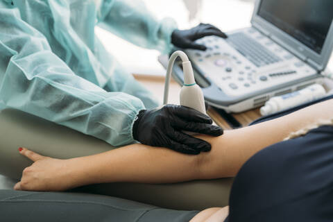 Arzt in Schutzkleidung untersucht den Arm einer Frau mit einem Ultraschallgerät, lizenzfreies Stockfoto
