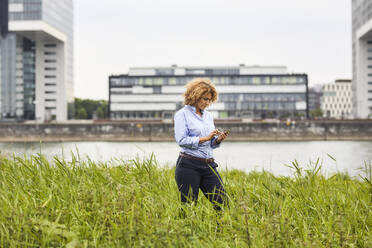 Businesswoman using smartphone at riverside in Cologne, Germany - MJFKF00330