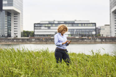 Geschäftsfrau mit Smartphone am Flussufer in Köln, Deutschland - MJFKF00330