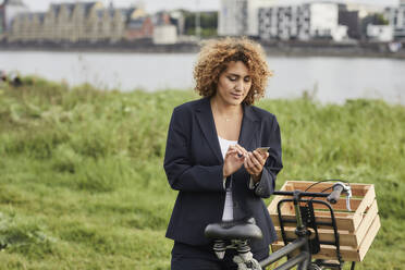 Businesswoman using smartphone at riverside in Cologne, Germany - MJFKF00339