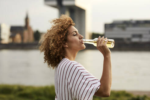 Porträt einer lächelnden Frau, die aus einer Glasflasche am Flussufer trinkt - MJFKF00342