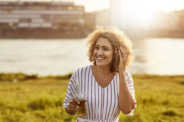 Porträt einer lächelnden Frau mit Glasflasche am Flussufer - MJFKF00346