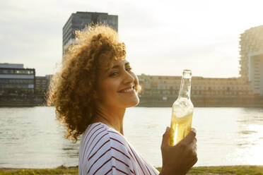 Porträt einer lächelnden Frau mit Glasflasche am Flussufer - MJFKF00352