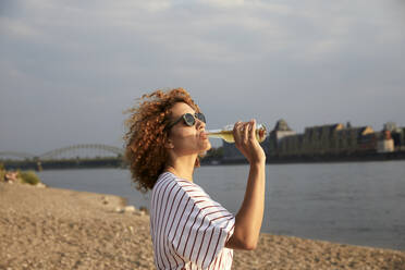 Porträt einer lächelnden Frau mit Glasflasche am Flussufer - MJFKF00355