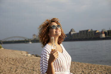 Portrait of smiling woman with glass bottle at riverside - MJFKF00356