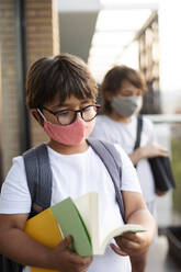 Siblings wearing masks outdoors, boy reading book - VABF03060