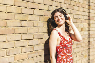 Glückliche junge Frau, die Musik hört und sich an eine Mauer lehnt - WPEF02993