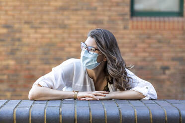 Young woman wearing protective mask looking sideways and leaning on wall - WPEF03010