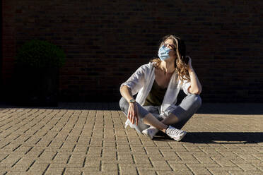 Young woman wearing protective mask, sitting on ground - WPEF03011