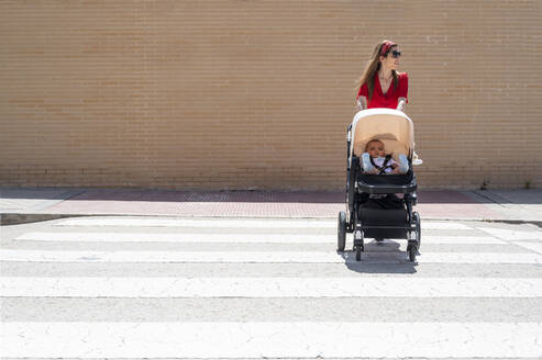 Mutter mit Sonnenbrille, die ihren Sohn im Kinderwagen schiebt, während sie auf der Straße spazieren geht - JCMF00798