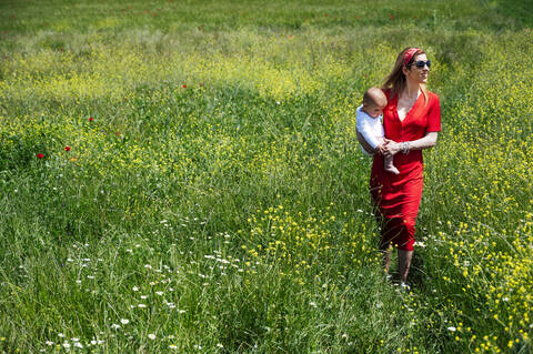 Frau trägt ihren kleinen Sohn bei einem Spaziergang auf einer Wiese an einem sonnigen Tag, lizenzfreies Stockfoto