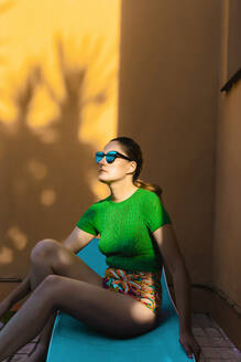 Frau mit Sonnenbrille sitzt auf Sonnenbank in bunten Hinterhof - ERRF04000
