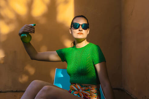Frau in farbenfroher Strandkleidung sitzt im Hinterhof und hält eine Sprühflasche - ERRF04001