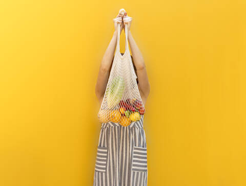 Junge Frau hält Früchte in einer Netztasche und steht vor einem gelben Hintergrund, lizenzfreies Stockfoto