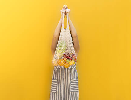 Young woman holding fruits in mesh bag while standing against yellow background - AFVF06607