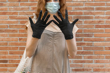 Woman wearing mask and gloves showing hands while standing against brick wall - AFVF06621