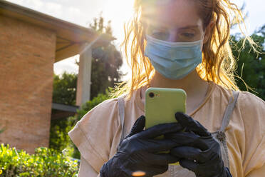 Nahaufnahme einer jungen Frau mit Maske, die im Hof ein Smartphone benutzt - AFVF06622