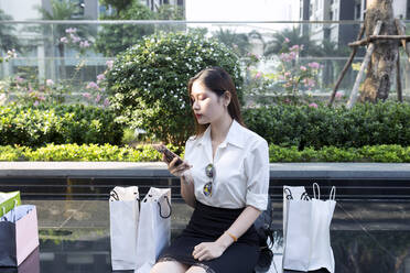 Junge Frau, die ein Smartphone benutzt, während sie mit Einkaufstüten auf dem Sitz in der Stadt sitzt - JPTF00517