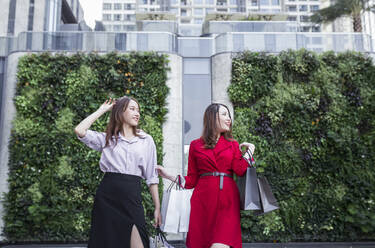 Smiling friends carrying shopping bags looking away while standing against building - JPTF00526