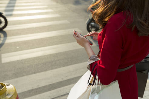 Young woman carrying shopping bags holding smart phone while crossing road in city - JPTF00534