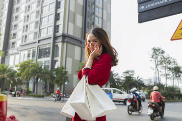 Smiling woman carrying shopping bags talking over smart phone while standing on street - JPTF00536