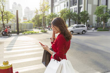Junge Frau trägt Einkaufstaschen und benutzt ihr Smartphone, während sie auf der Straße steht - JPTF00537