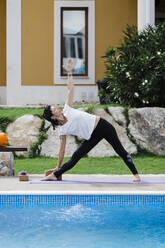 Woman practicing triangle position at poolside against house in garden - DCRF00274