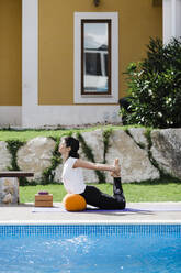 Woman practicing bow pose while leaning on bolster at poolside in yard - DCRF00277