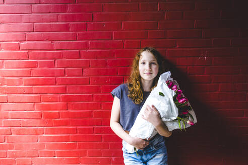 Girl holding bouquet while standing against red wall in city - IHF00340