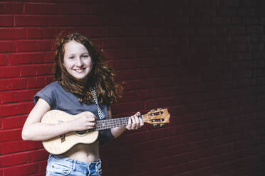 Lächelndes Mädchen spielt Ukulele, während sie gegen eine Backsteinmauer in der Stadt steht - IHF00341