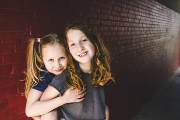 Cute girl embracing sister while standing against brick wall - IHF00342