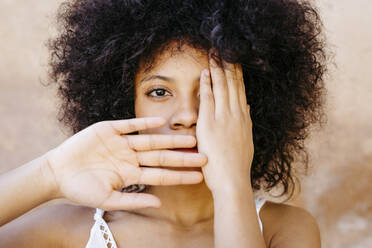 Schwarze Frau, die vor einer Wand steht und ein Auge und den Mund mit den Händen bedeckt - TCEF00735
