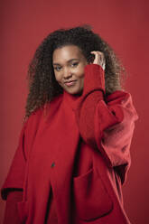 Lächelnde Frau mit lockigem Haar, die einen Mantel trägt und vor einem roten Hintergrund steht - SNF00312
