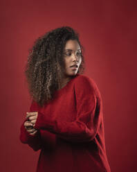 Serious thoughtful woman with curly hair standing against red background - SNF00314