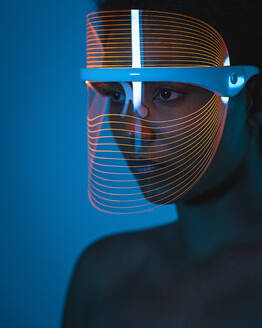 Nahaufnahme einer Frau, die eine Blaulichttherapie-Maske trägt und zu Hause an der Wand lehnt - SNF00325