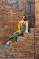 Selbstbewusste junge Frau, die auf einer Treppe vor einer Backsteinmauer sitzt - AFVF06640