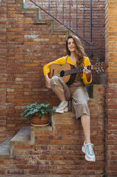 Lächelnde junge Frau, die auf einer Treppe sitzend Gitarre spielt - AFVF06645