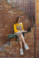 Junge Frau mit langen Haaren, die einen Laptop benutzt, während sie auf einer Treppe vor einer Backsteinmauer sitzt - AFVF06647