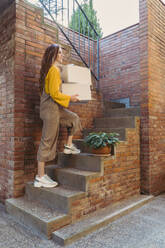 Junge Frau, die weiße Kisten trägt, während sie eine Treppe an einer Backsteinmauer hinaufgeht - AFVF06655
