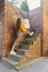 Verspielte junge Frau, die Kisten trägt, während sie eine Treppe an einer Backsteinmauer hinaufgeht - AFVF06656