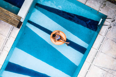 Aerial View Of Tanned Woman Floating Comfortably On Orange Inflatable Blow Up Ring In Luxury Villa Swimming Pool - AAEF08912