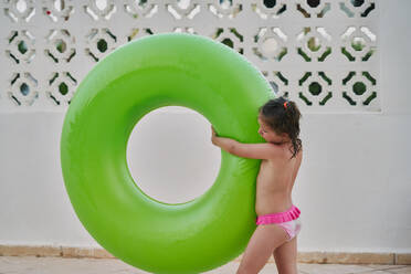 Girl carries a green float in her arms by the pool - CAVF84608