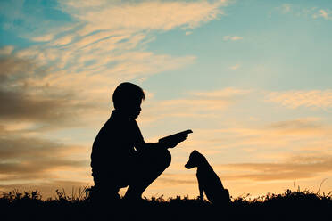 Silhouette Junge liest Buch und sitzt bei Welpen gegen bewölkten Himmel bei Sonnenuntergang - EYF05171