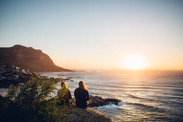 Rear View Of Female Friends Sitting At Beach Against Clear Sky During Sunset - EYF05227