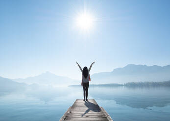 Full Length Rear View Of Woman With Arms Raised Standing On Pier By Lake Against Sky During Sunny Day - EYF05394