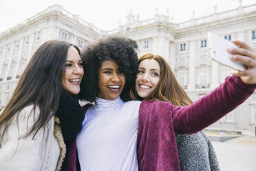 Happy young woman taking selfie with female friends while standing against Madrid Royal Palace, Spain - ABZF03177