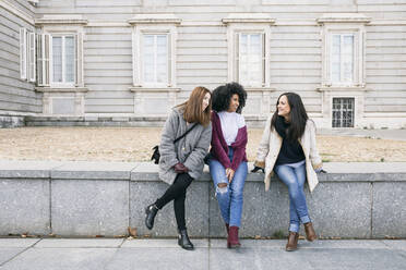 Fröhliche Freundinnen unterhalten sich, während sie auf einer Mauer vor dem Königspalast in Madrid sitzen, Spanien - ABZF03179