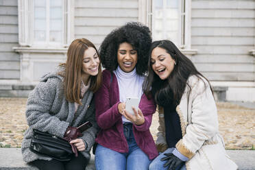 Cheerful female friends using smart phone while sitting against Madrid Royal Palace, Spain - ABZF03185