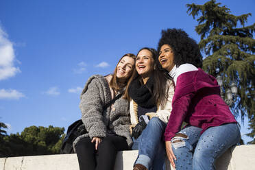 Fröhliche Freundinnen schauen weg, während sie auf einer Stützmauer gegen den blauen Himmel im Park sitzen - ABZF03190